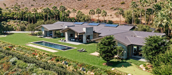 A stretch of a home with solar panels on top, a rectangular pool with the desert landscape in the background