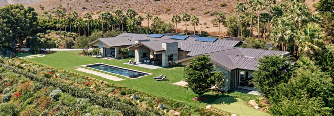 A stretch of a home with solar panels on top, a rectangular pool with the desert landscape in the background