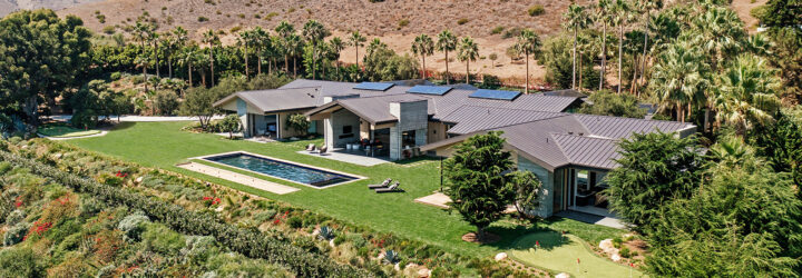 A stretch of a home with solar panels on top, a rectangular pool with the desert landscape in the background