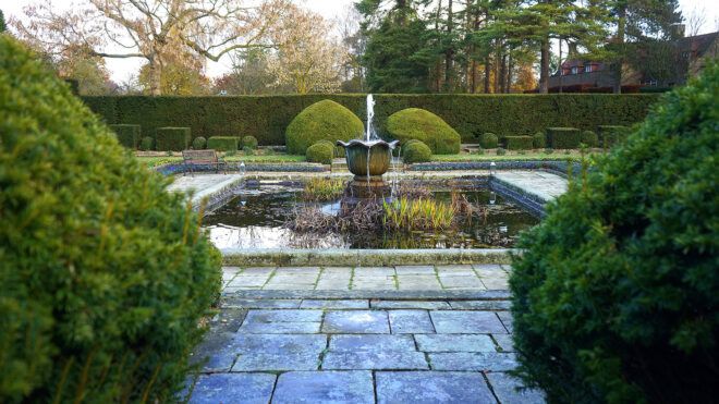 A square pond with a fountain in the center of it within a garden