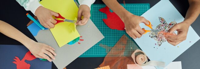 Five hands surrounding a table cluttered with papers and crafting