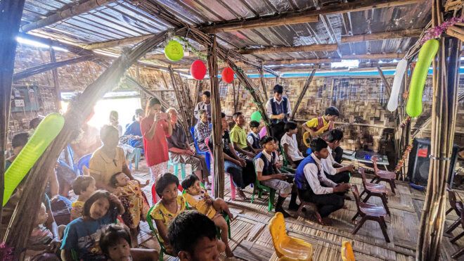 An interior of a bamboo structure filled with people sitting and standing facing the front