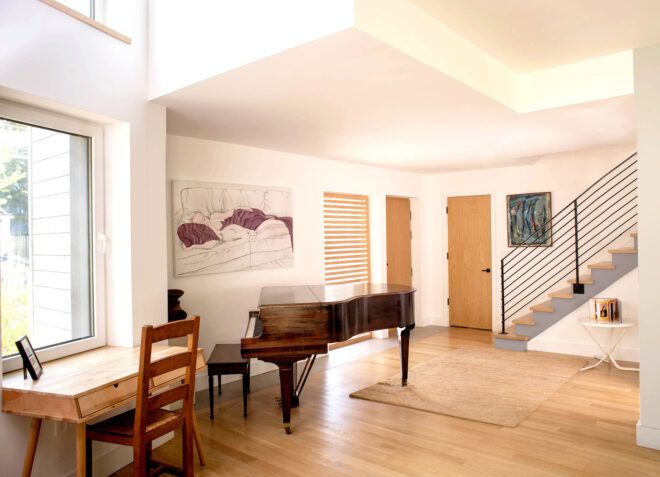 A piano sitting in a living room space with stairs leading up to the second floor and a desk facing a window
